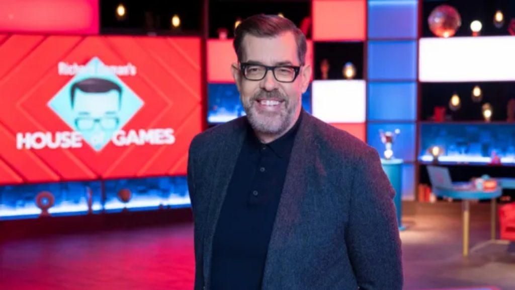 Richard Osman wearing a navy suit and glasses on the House of Games set