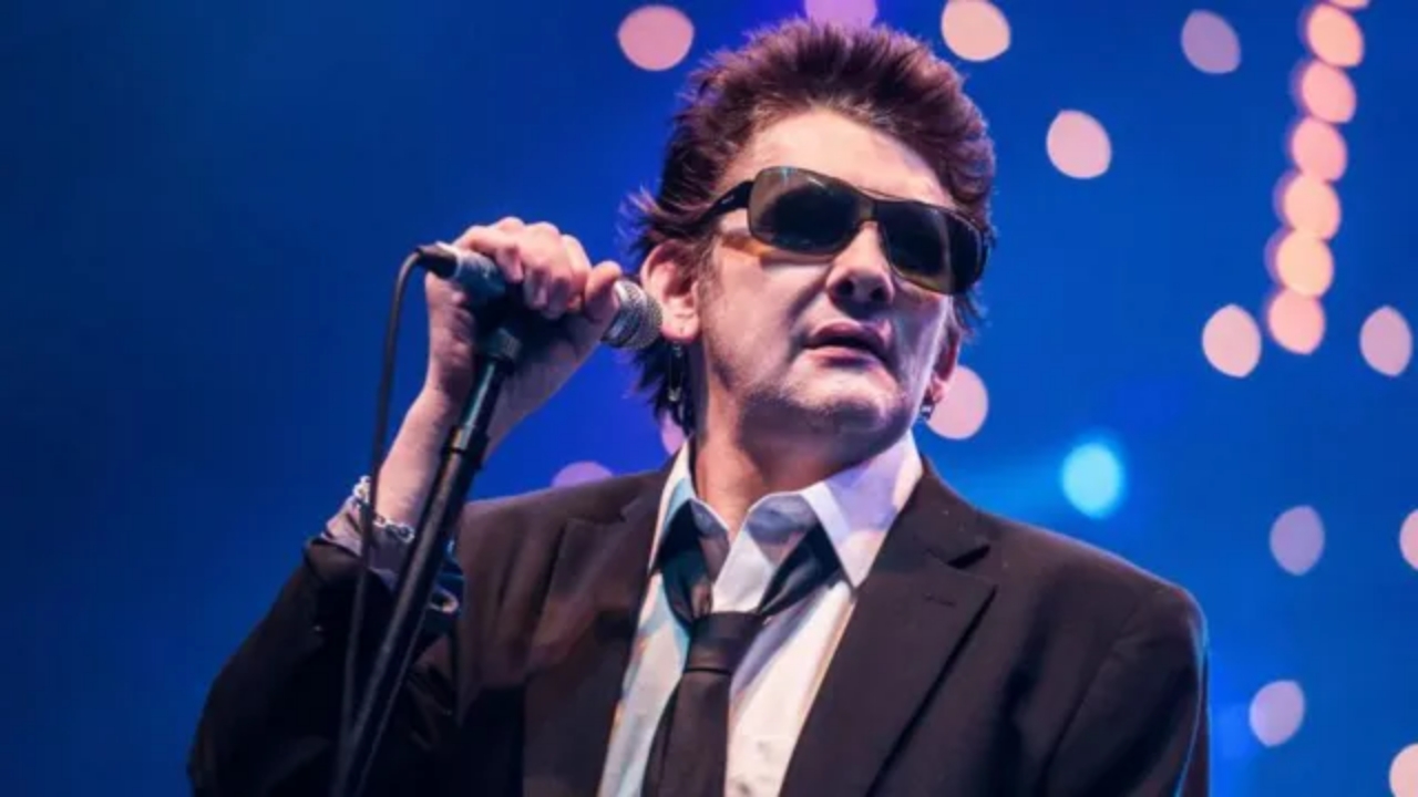 Shane MacGowan performing on stage holding a microphone during a live concert.
